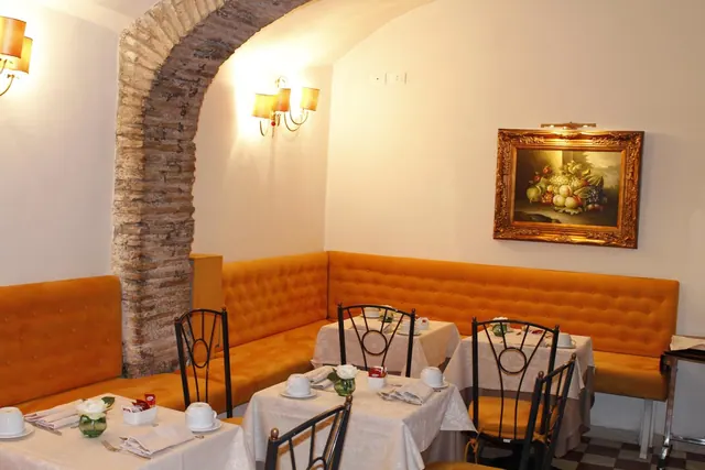 A dining room with set tables, orange tufted banquette seating, a brick archway and a framed still life painting on the wall.