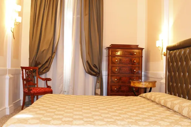 A bedroom with a made bed under a tufted leather headboard, a red armchair, a wooden chest of drawers, a small round side table, wall lights and tall curtains.