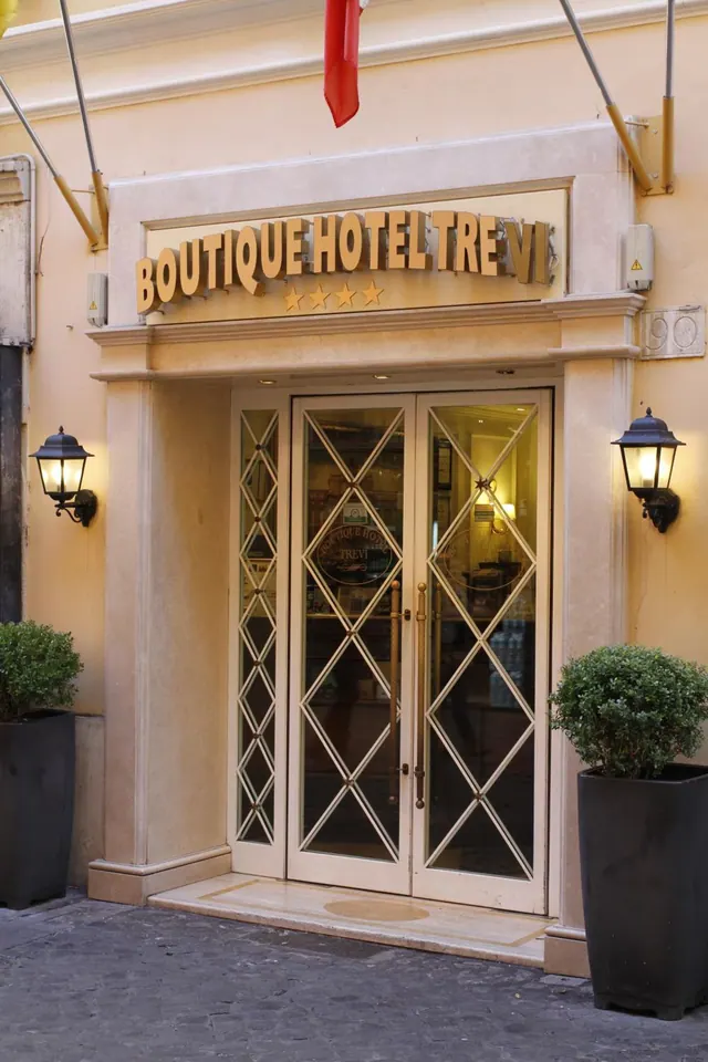 The entrance of Boutique Hotel Trevi, with a lit sign above glass double doors, wall lanterns on each side, and potted topiary on the cobbled pavement.