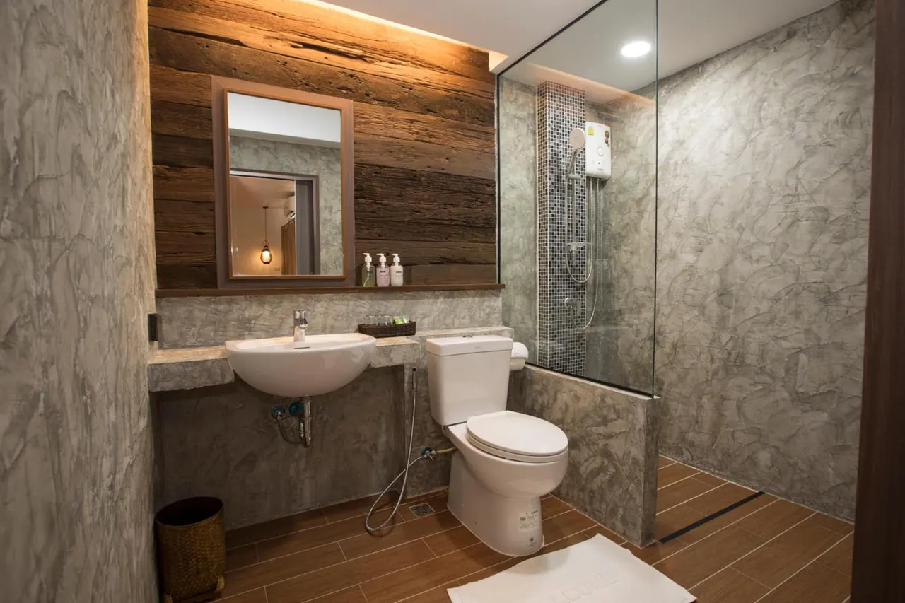 A bathroom with a wall-mounted basin, a toilet with a handheld bidet sprayer, a glass-enclosed walk-in shower, and a wooden plank accent wall beside a mirror and toiletry bottles.