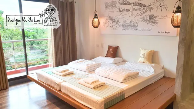 A bedroom with a wooden platform bed holding two mattresses, white bedding, pillows and folded towels, with a sliding door to a balcony and green trees outside.