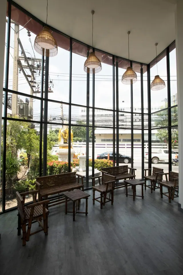 A glass-walled seating area with bamboo chairs and tables and woven pendant lamps overlooks a street with trees, parked cars and a golden spirit house.