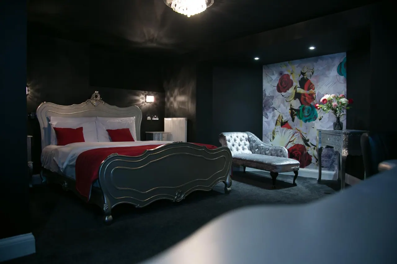 A dark bedroom with an ornate silver bed, red bedding and throw, a tufted chaise longue, and a floral mural on the wall.