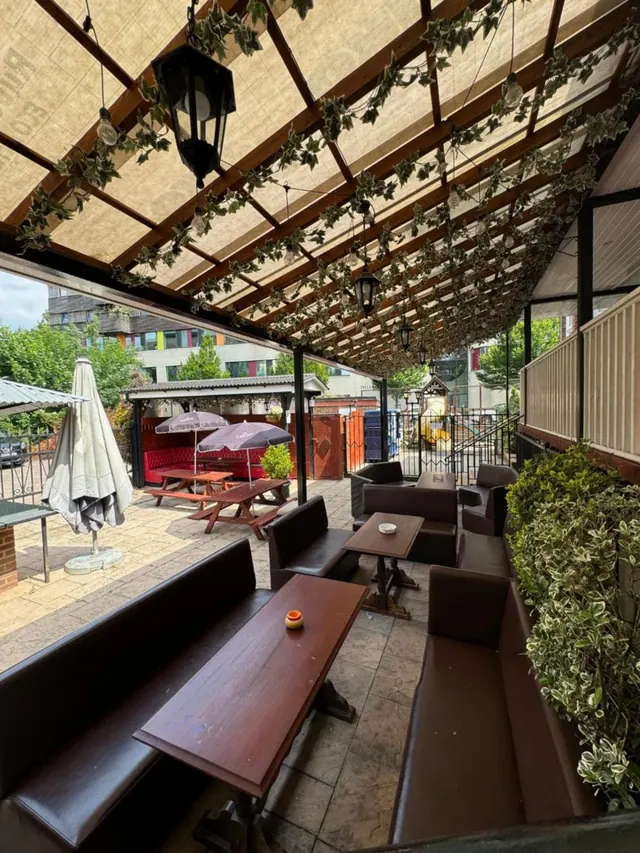 A covered outdoor terrace with an ivy-draped pergola, wooden tables, leather banquettes and parasols on a paved patio.