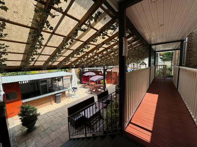 A covered decking terrace with white railings and a pergola overlooks a paved courtyard with picnic benches and parasol umbrellas.