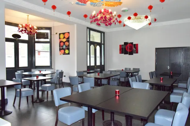 An empty dining room with dark tables, light blue upholstered chairs, red paper lanterns and two chandeliers hanging from a decorated ceiling.