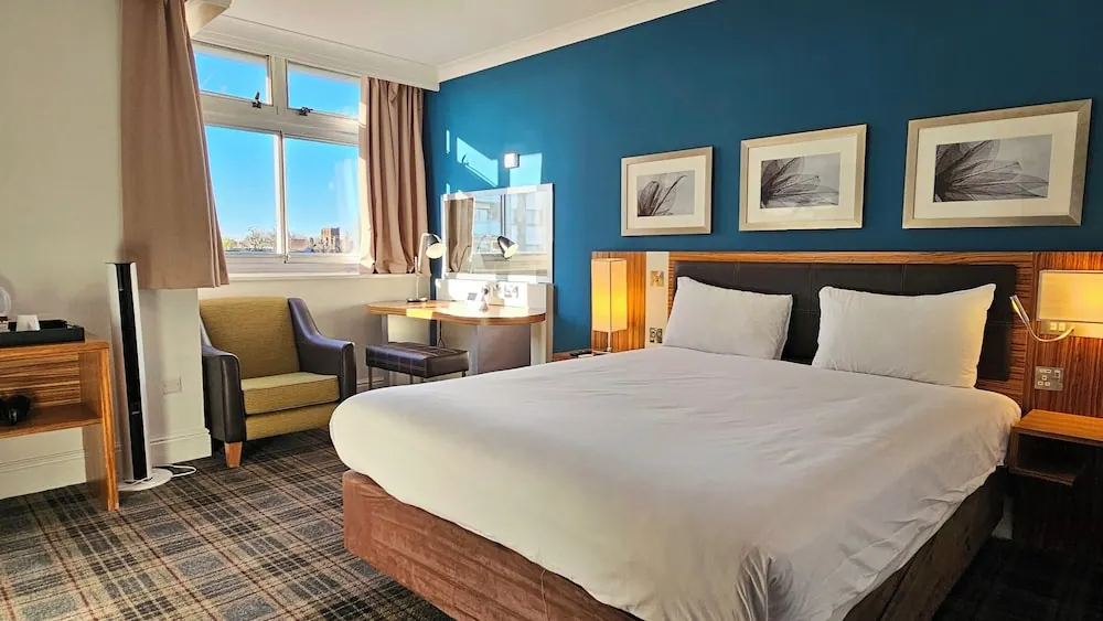 A double bed with white bedding sits against a dark blue accent wall, beside an armchair, a desk with a mirror and a window showing buildings outside.