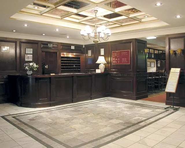 A hotel lobby with a dark wood reception counter, a coffered ceiling with a chandelier, and a marble floor with a patterned border.