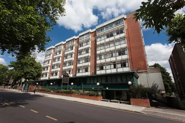 A multi-storey brick and white-panelled hotel building with a dark green ground floor stands beside a tree-lined street under a partly cloudy sky.