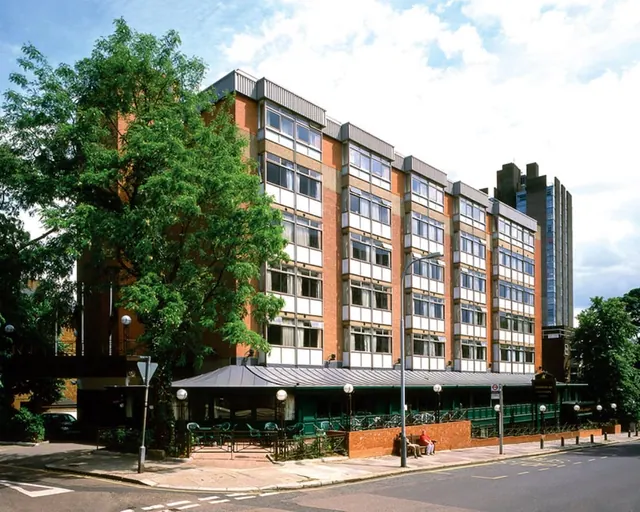 Exterior view of a multi-storey brick hotel building with white-paneled windows, a green canopy over outdoor tables and chairs, and a large tree on the left.