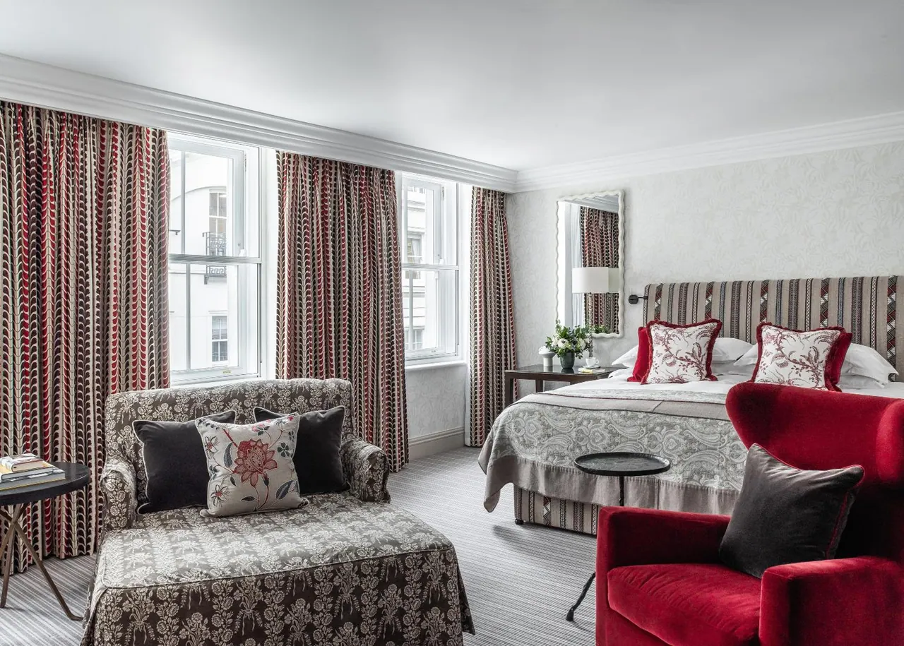 A hotel bedroom with a made double bed, a patterned sofa and a red armchair, with tall windows hung with striped curtains.