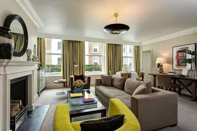 A furnished living room with a grey sofa, yellow armchair, glass coffee table, fireplace and a desk by the windows.