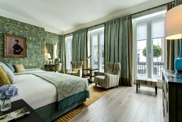 A bedroom with a large bed, striped armchairs and tall windows looking out onto a street with a building opposite.