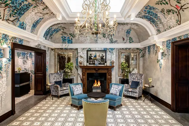 A lobby seating area with a crystal chandelier, a wisteria-patterned mural on the walls and ceiling, a fireplace, and blue and patterned armchairs on a geometric tiled floor.