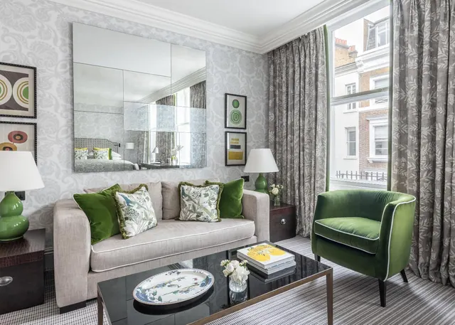 A living area with a grey sofa, a green velvet armchair, and a large mirror on a patterned wall beside a tall window showing buildings across the street.