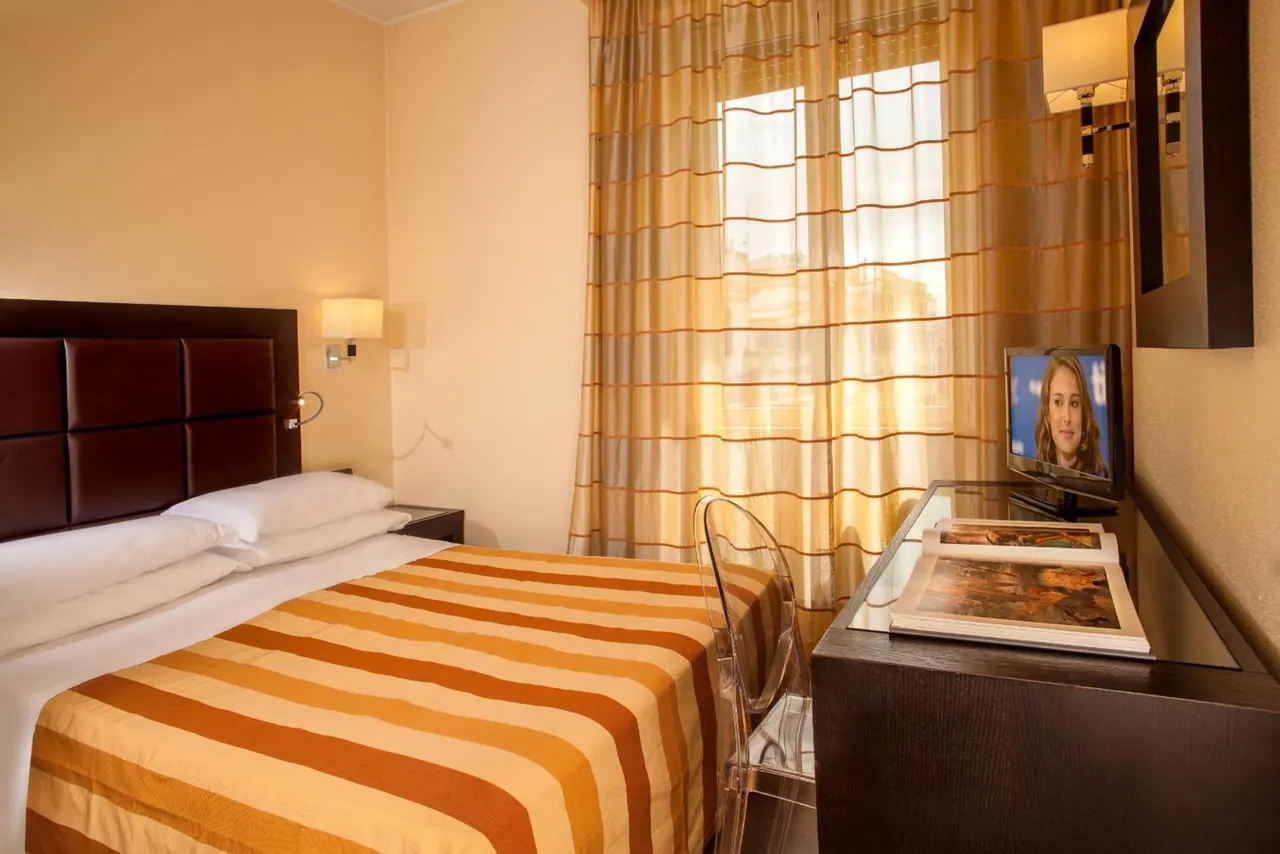 A hotel bedroom with a double bed under a striped orange throw, a glass desk holding books and a flat-screen TV, and striped curtains over the window.