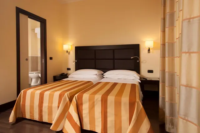 A hotel bedroom with two beds placed side by side under a dark padded headboard, with an open sliding door showing a toilet in the en suite bathroom.