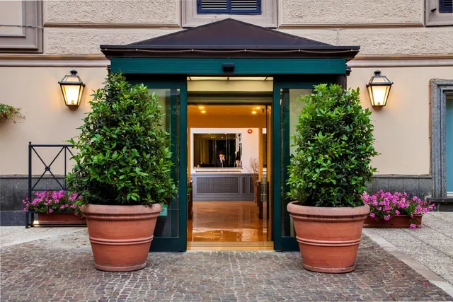 Entrance of a hotel with glass doors open onto a lit reception desk, flanked by two large potted shrubs, pink flowering plants and wall lanterns.