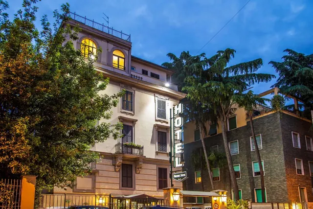 Evening exterior of a cream-coloured hotel building with lit arched windows and a vertical HOTEL sign, beside a dark brick building and tall trees.