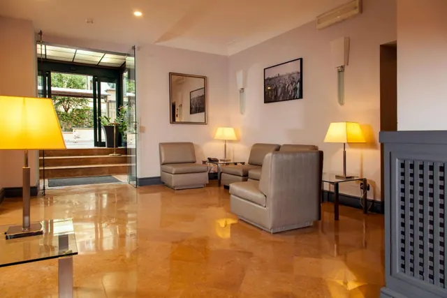 A lobby seating area with leather armchairs, table lamps, framed prints and a polished stone floor, with a glass entrance door and steps leading outside.