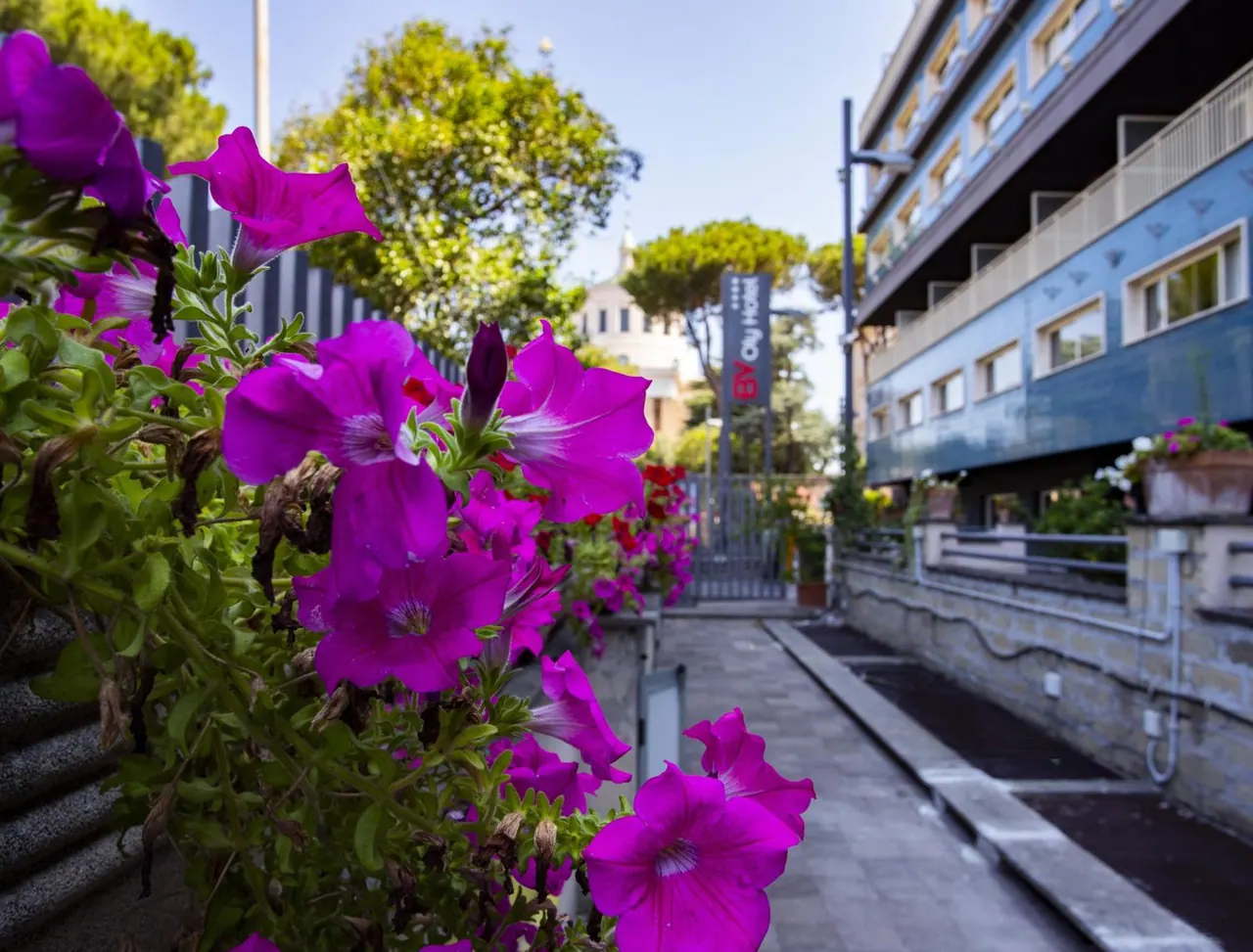 A pedestrian passage lined with pink petunias in planters runs past a blue hotel building toward a gate with a hotel sign.