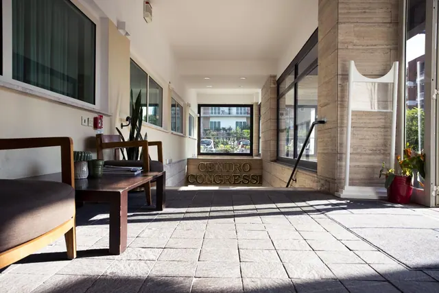 A covered entrance area with a wooden bench, a potted plant, and a stone sign reading Centro Congressi, with a view through glass to a building and parked cars.