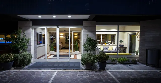 Night view of the hotel entrance with glass sliding doors, potted plants, and a lit lounge with a grey sofa visible through the window on the right.