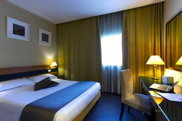 A hotel bedroom with a double bed, bedside lamp, framed prints, and a desk with a laptop and lamp beside a curtained window.