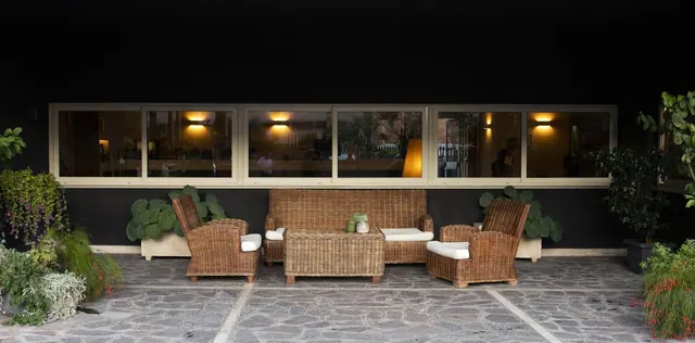 An outdoor terrace with two wicker armchairs, a wicker sofa and a low wicker coffee table on a stone-paved floor, with lit windows of the building behind.