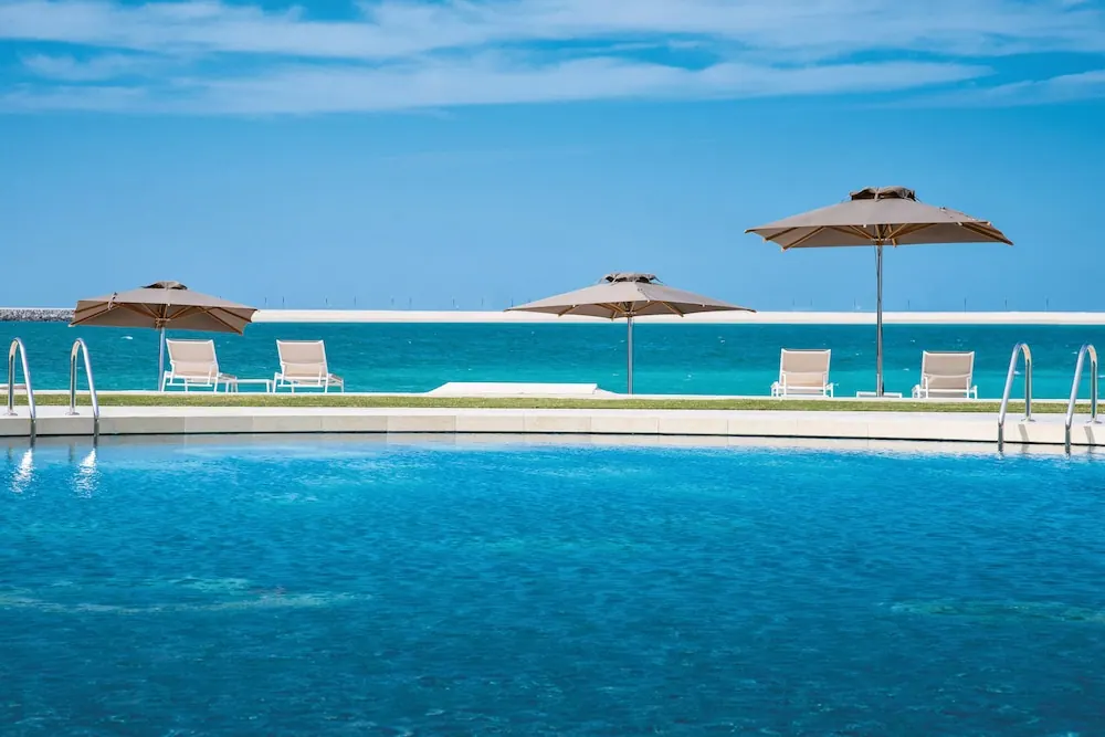 A large swimming pool with sun loungers and shaded umbrellas beside it, with the sea visible beyond.