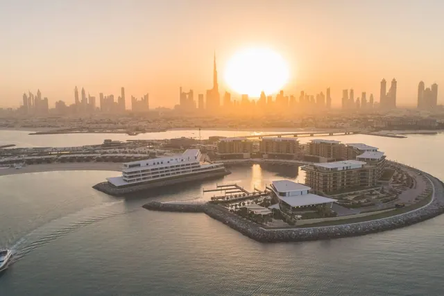 Aerial view of a waterfront hotel complex on a peninsula at sunset, with a city skyline including the Burj Khalifa in the background.