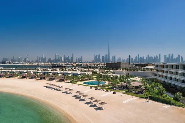 Aerial view of a sandy beach with rows of shaded umbrellas and sun loungers, a round pool, low villas and a hotel building, with the Dubai skyline including Burj Khalifa in the distance.