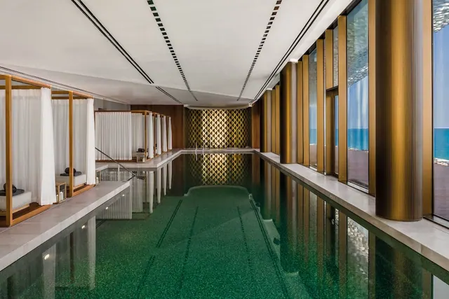 An indoor lap pool with green tiles, white-curtained cabana beds along one side, and floor-to-ceiling windows looking out onto the sea.