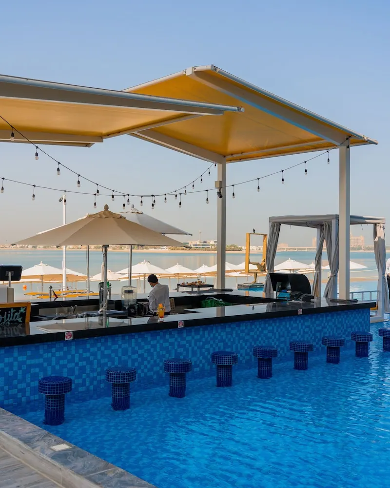A swim-up pool bar with blue tiled stools in the water, a bartender working behind the counter, and white umbrellas on a beach with the sea beyond.