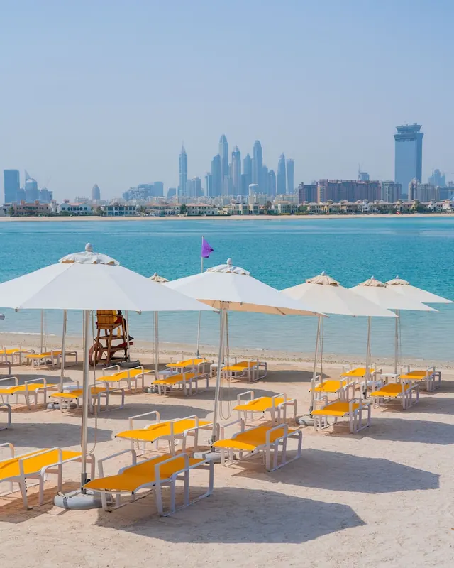 Rows of white parasols and yellow sun loungers sit on a sandy beach facing turquoise sea and a city skyline of high-rise towers.