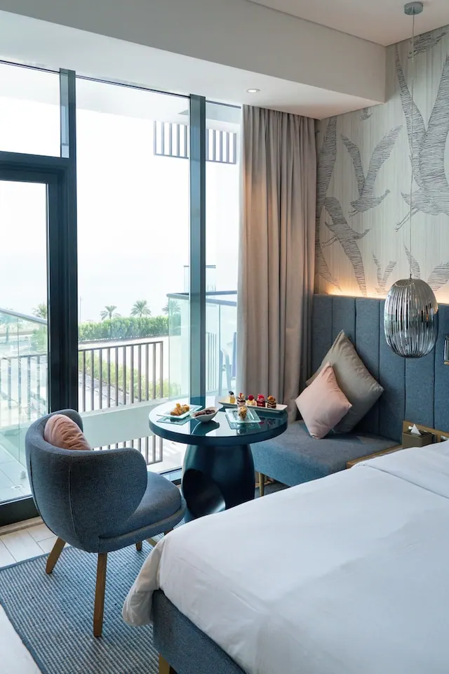 A hotel bedroom with a large bed, a round table and armchair by floor-to-ceiling windows, with a balcony and palm trees visible outside.