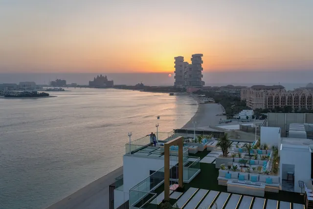 Sunset over the sea seen from a rooftop terrace with white lounge seating, with the Atlantis hotel visible in the distance and two people standing at the glass railing.