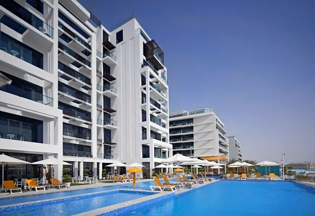 A large outdoor swimming pool with orange sun loungers and white umbrellas lies in front of a white multi-storey building with balconies under a clear blue sky.