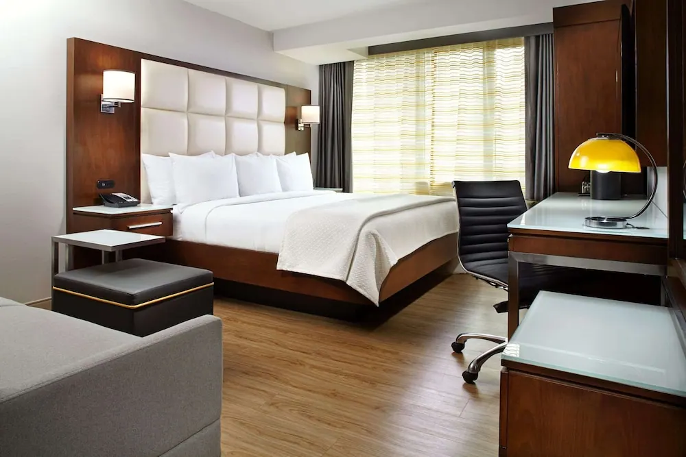 A bedroom with a king bed under a white quilted headboard, a wooden desk with a yellow lamp and a grey sofa in the foreground.