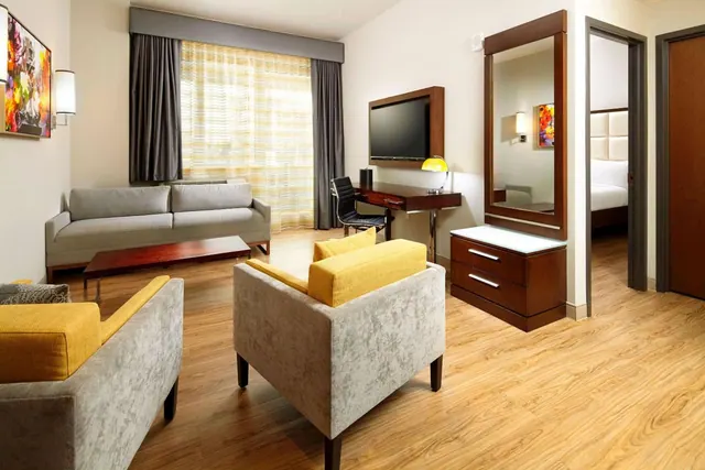 A hotel suite living area with a grey sofa, yellow armchairs, a wall-mounted television, a desk and a wooden dresser, with a bed visible through an open doorway.