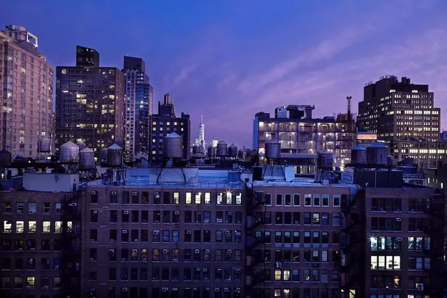 A twilight view over rooftops of lit buildings and water towers, with One World Trade Center visible in the distance.