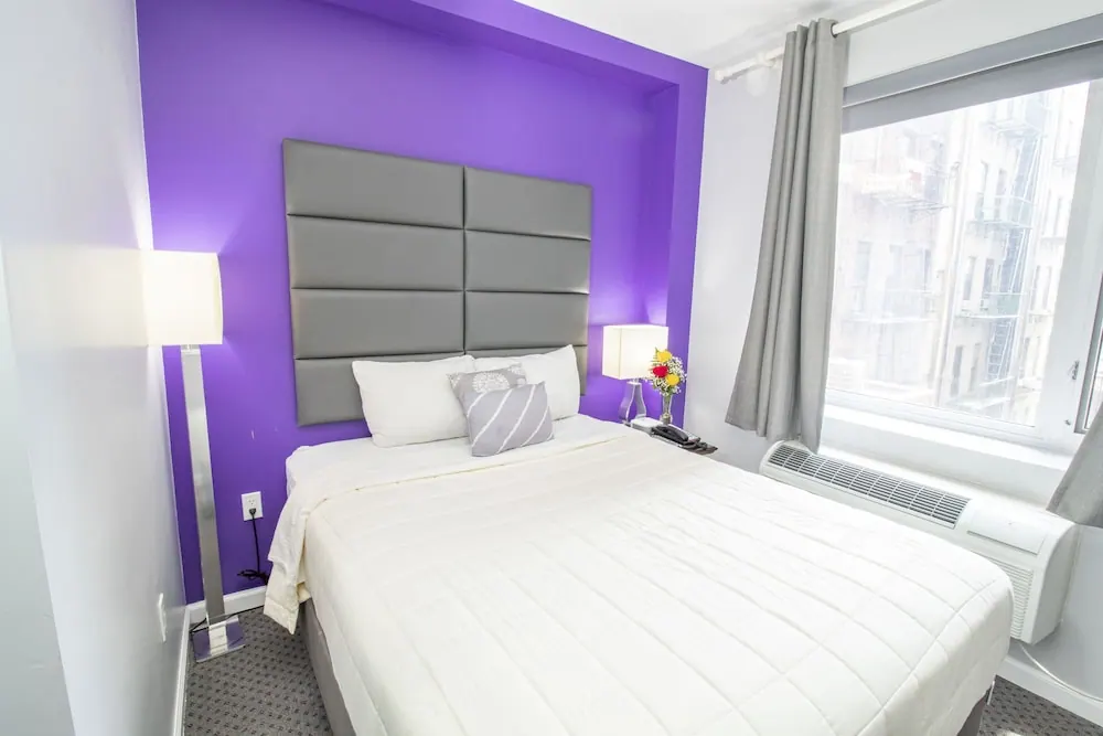 A bedroom with a purple accent wall, a grey upholstered headboard, a white bed, and a window looking out at a neighbouring building with fire escapes.