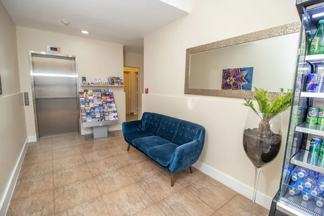 A lobby seating area with a blue velvet sofa, a framed mirror, a potted plant in a tall glass vase, a brochure rack, a lift and a glass-door drinks fridge.