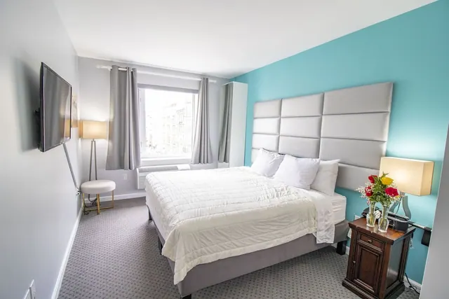 A hotel bedroom with a king bed, a teal accent wall, a wall-mounted television, and a window looking out onto neighbouring buildings.