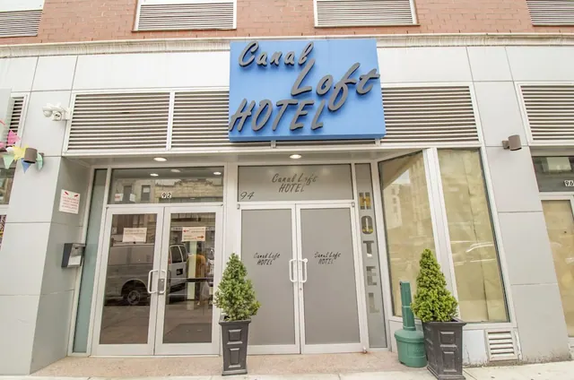 The street entrance of the Canal Loft Hotel, with a blue sign above glass double doors and two potted topiary plants on the sidewalk.