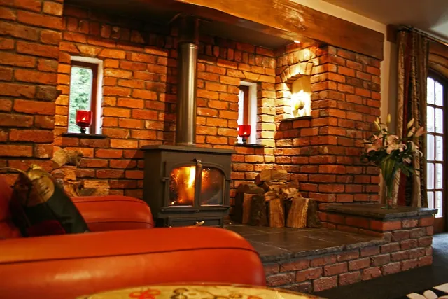 A living room with a lit wood-burning stove set in a exposed brick wall, an orange leather sofa in the foreground, stacked firewood on the stone hearth, and a vase of lilies by the tall window.