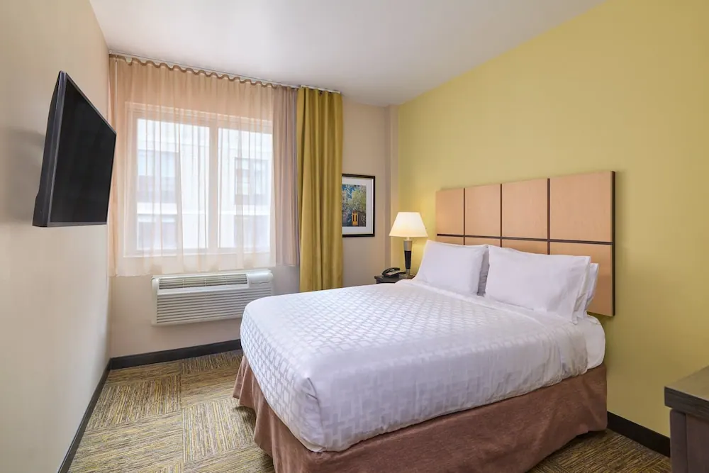 A bedroom with a made double bed, a wall-mounted television, a window with sheer curtains and a wall air conditioner unit.