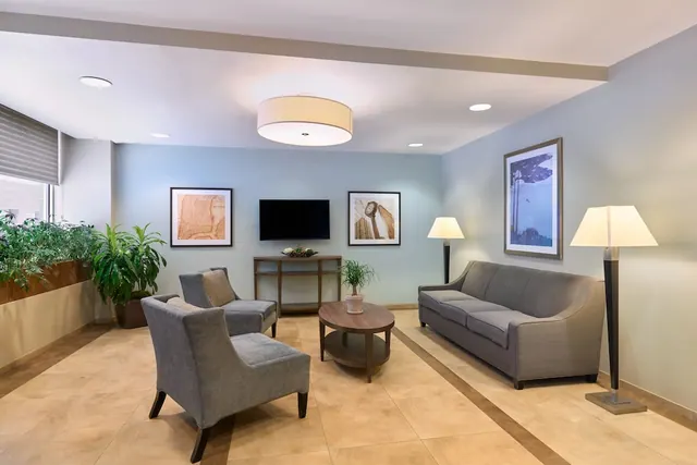 A lounge seating area with two grey armchairs, a grey sofa, a round wooden coffee table, floor lamps, potted plants and a wall-mounted television.