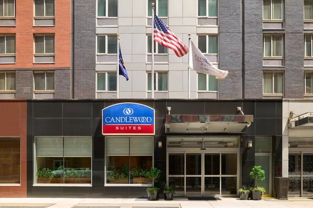 The front of a Candlewood Suites building shows its blue and red sign, a glass entrance with a canopy, flags on poles above, and potted plants at street level.