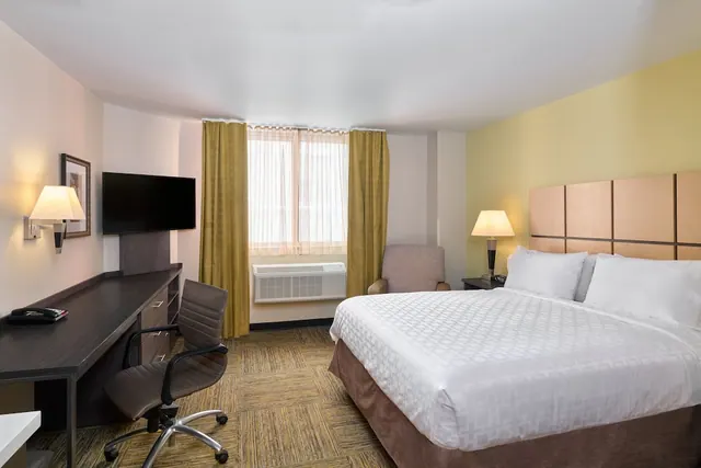 A bedroom with a king bed, a desk with a chair, an armchair, a wall-mounted television and a window air conditioner under the window.